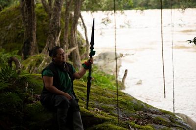 A woman sits beside a river holding a spear.