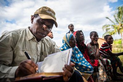 A man writes in a notebook as several other people look on in the background.