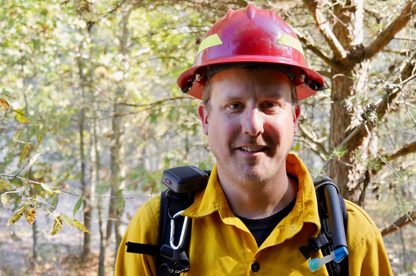 Gabe Cahalan headshot. A smiling man wearing yellow protective fire gear and a red hardhat stands in a forest during a controlled burn.