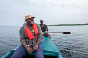 Bowie Osstantou smiles and laughs while sitting in a narrow turquoise colored fishing boat in the middle of a lake. A fisherman sites behind him on the edge of the boat.