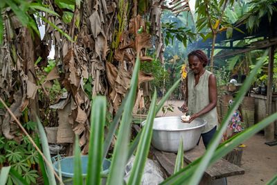 a woman smiles while cutting up stew ingredients over a large aluminum pot in an outdoor kitchen area surrounded by tropical trees and plants.