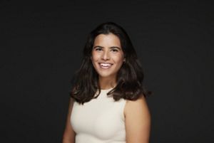 A headshot of a woman with shoulder length dark brown hair wearing a cream colored sleeveless top in front of a dark background.