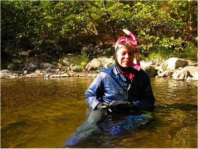 A woman sits in a shallow creek, wearing a wetsuit and pink scuba gear. 