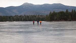 Three people cross a frozen pond with mountains in the background.