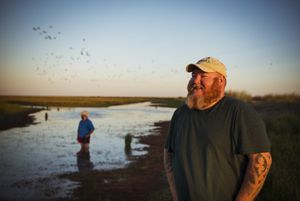 A man smiles while standing in a marsh with another man and flocks of flying birds in the background.