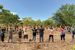 A large group of people gathered outdoors in a circle with their arms raised during an Olowalu work day.
