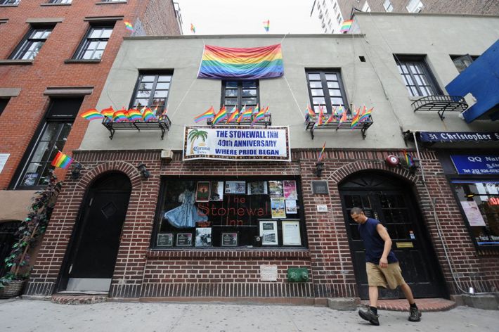 A man walks passed the urban business storefront of NY's Stonewall Inn. Two arched doorways flank a wide picture window. The tall windows above the entrance are overhung with a rainbow Pride banner.