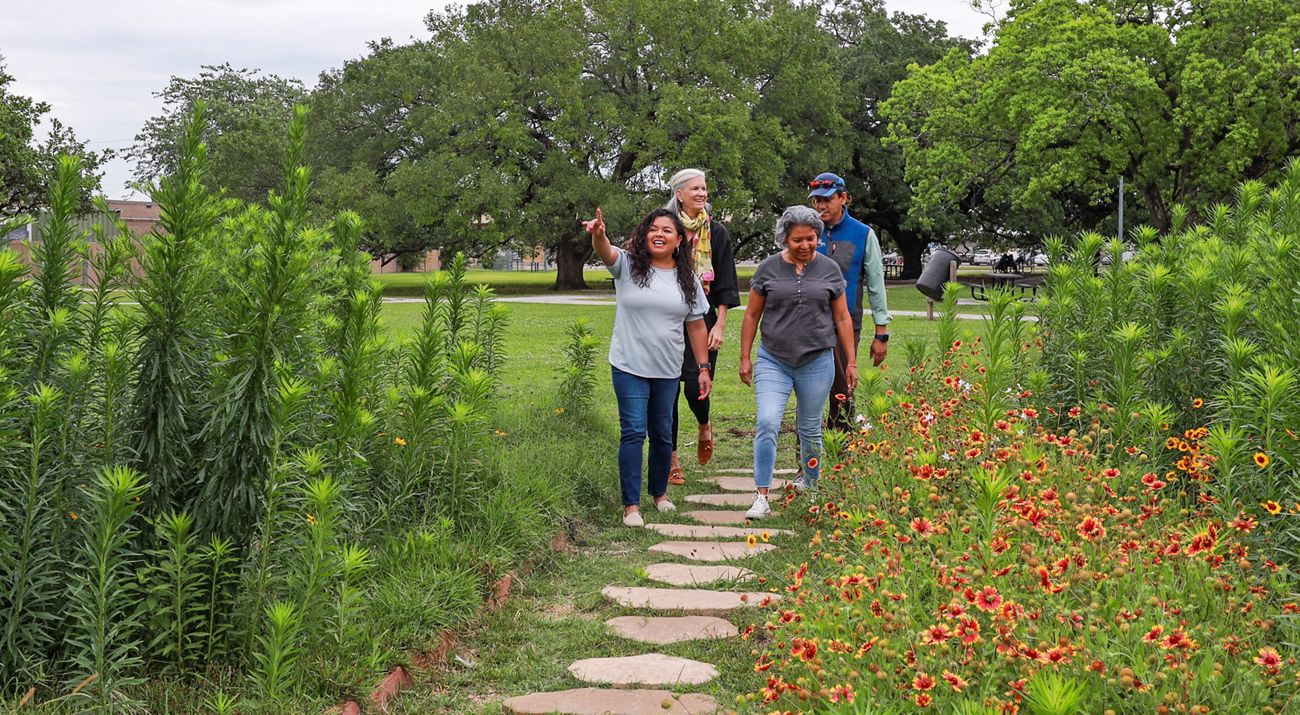 Three women and one man walk on a path with flowers in a green park.