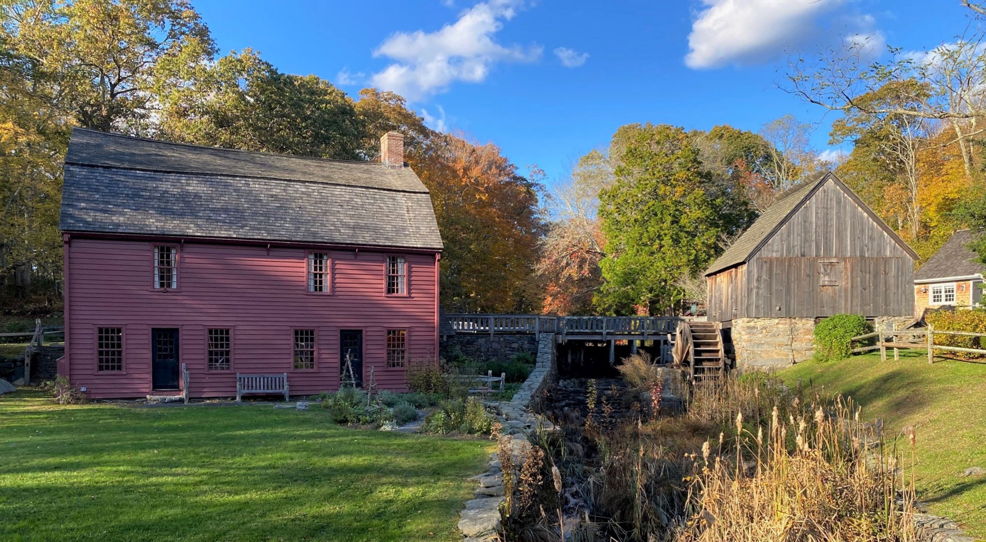An 18th century home, painted deep red, sits beside a stone mill race and small, wooden grist mill. 
