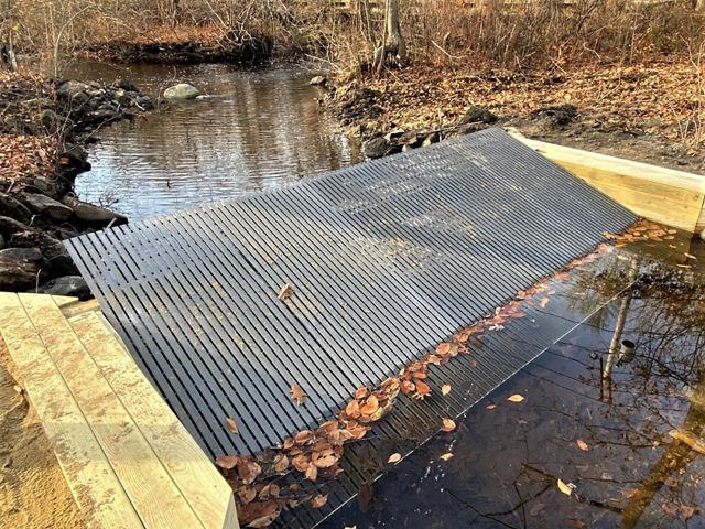Gray fiberglass grating rests at an angle on wooden abutments in a small, calm stream.