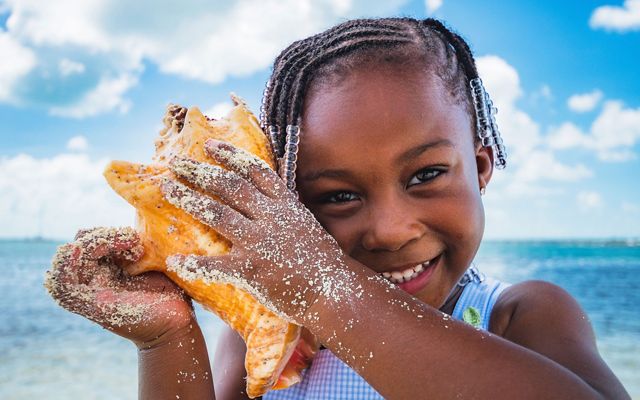 A small child holds a conch up to her ear and smiles for the camera.