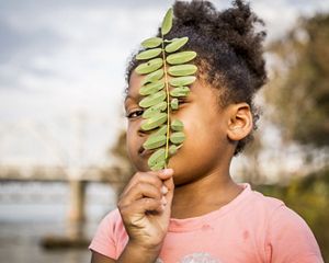 una niña sostiene una pequeña rama frente a su rostro y coloca su mirada entre los espacios de las hojas.
