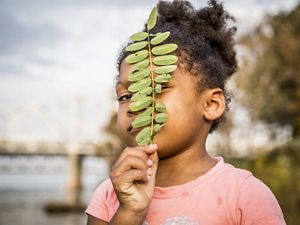 una niña sostiene una pequeña rama frente a su rostro y coloca su mirada entre los espacios de las hojas.