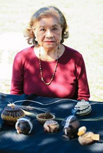 A woman sits at a table with baskets and animals made of pine needles.