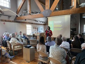 People sit in rows of chairs in an room with high ceilings and wood beams, listening to a presenter at the front of the room who stands in front of a projector screen. 