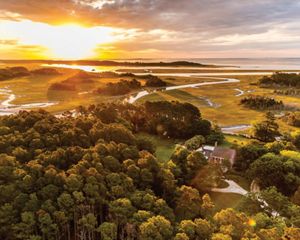 Aerial view of Brownsville Preserve. An historic brick home with a circular oyster shell driveway stands in a clearing between thick stands of trees. In the background the sun rises over the Atlantic.