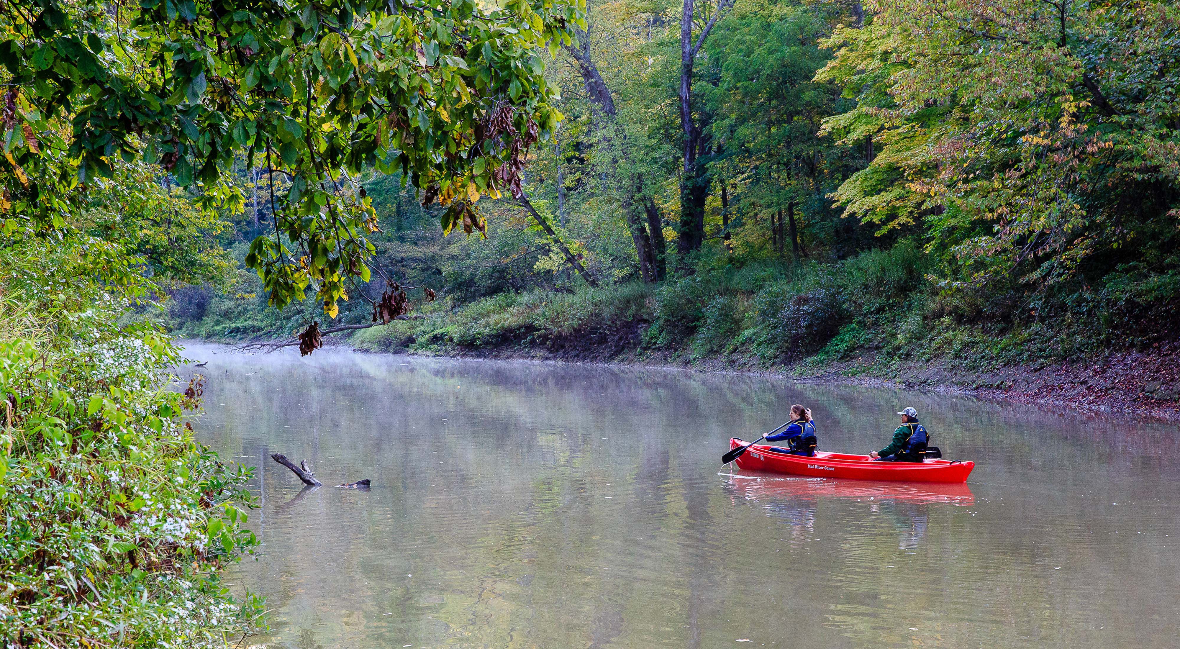 Two people paddle a red plastic canoe on a calm, broad stream lined with trees.