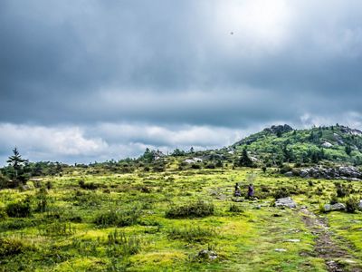 Two people follow a curving, rocky dirt path through an open field approaching a forested mountain ridge.
