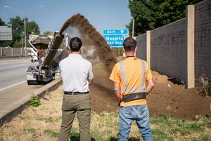 Two men stand together watching soil being spread from the back of a dump truck onto a planting area tucked between a highway and tall sound barrier wall.