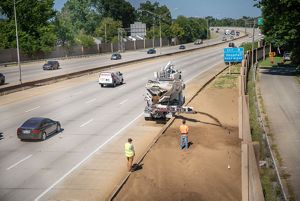 Two people watch as soil is distributed onto a planting area adjacent to a highway as cars pass by.