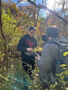 Seequan Weeden stands surrounded by trees and branches, holding a tray of bulrush seedlings in one hand and using the other to hold up one clump of grass for podcast producer Jay Feinstein.