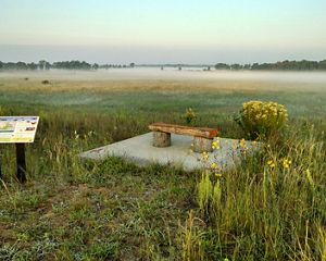 Landscape view overlooking a wide, flat prairie with a layer of fog over it in the distance and a wood-and-stone bench and interpretive sign in the foreground.