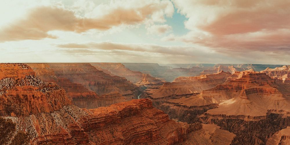 Bright golden sun rays fall over the red-colored Grand Canyon under a mostly cloudy sky.