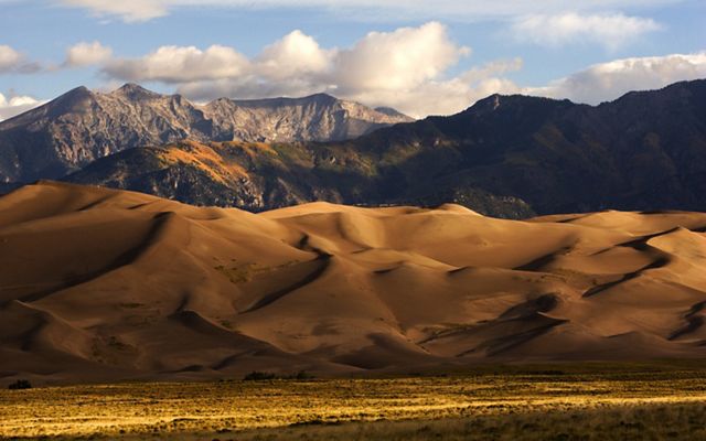 A sunset landscape of Great Sand Dunes National Park, with golden dunes in the foreground and layers of rugged mountains in the background. 