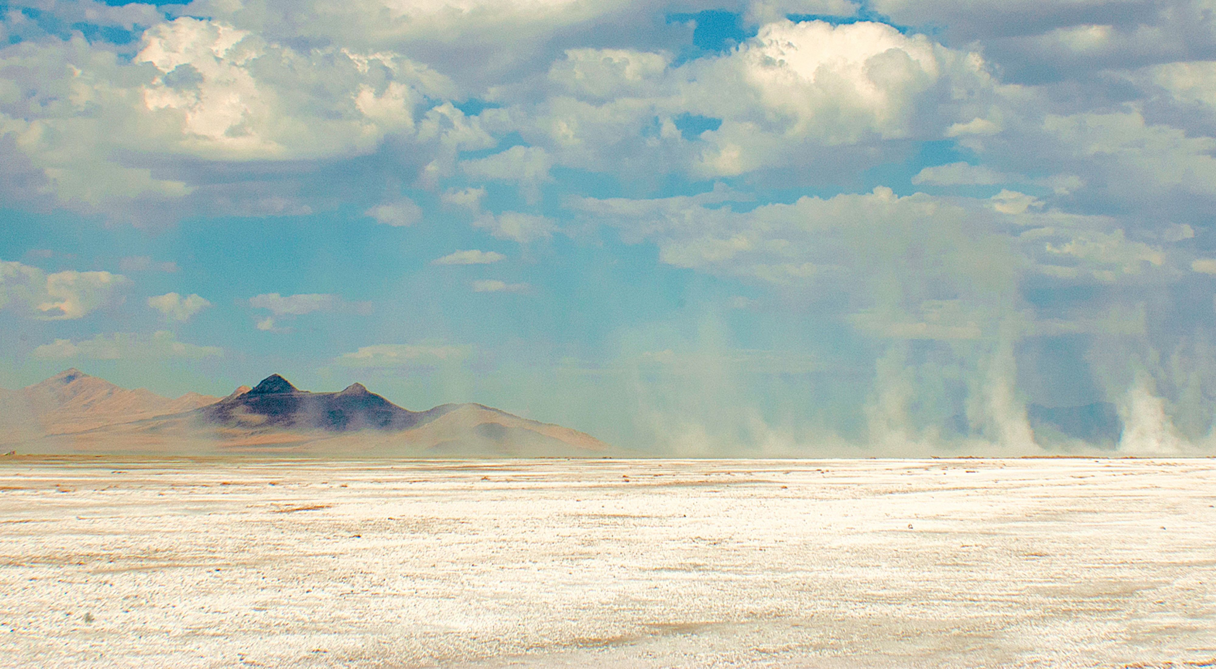 Dust and sand blown by wind across a white dry lakebed under cloudy blue skies with a dark mountain in the background. 