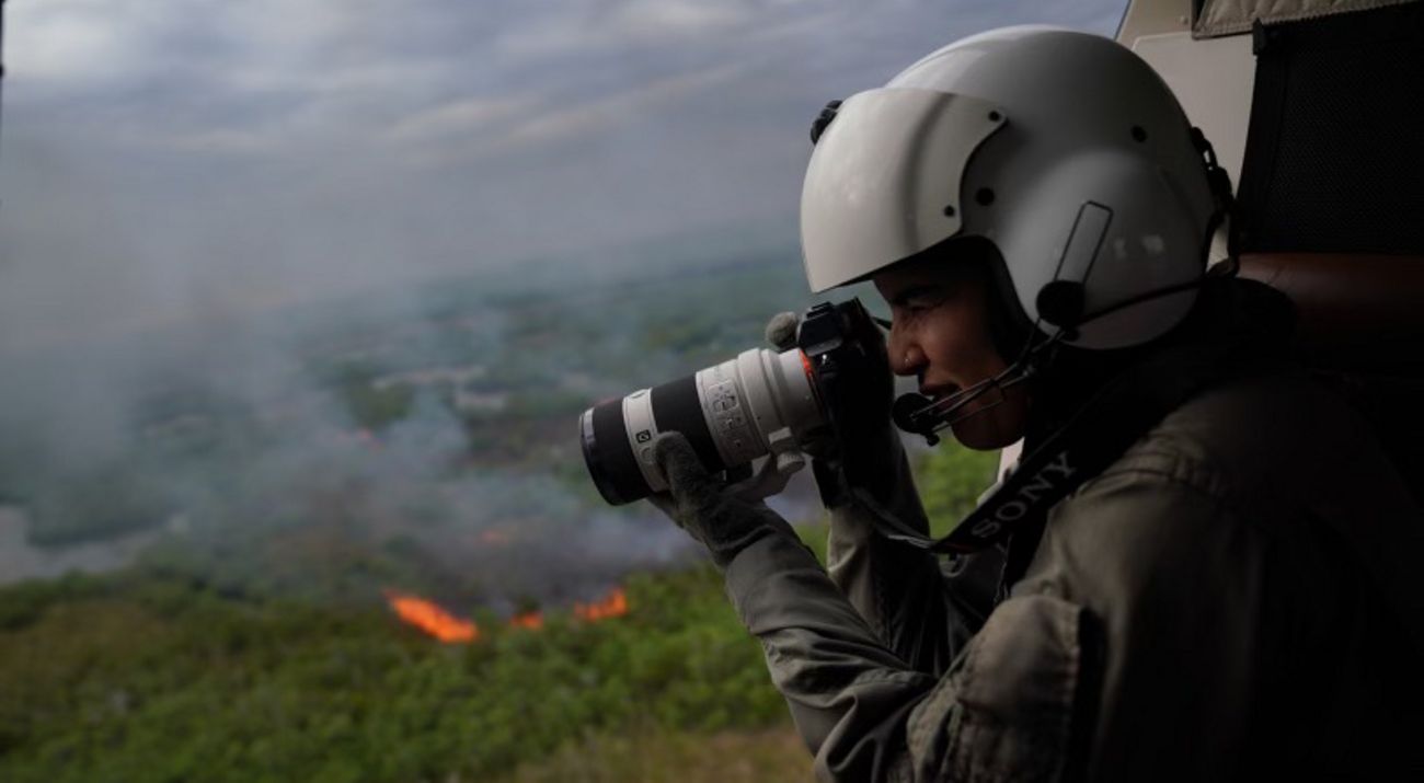 A woman in a helicopter holding a camera, taking photos of a prescribed burn beneath her.