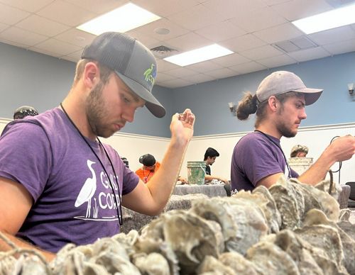 Two men wearing gray hats and purple shirts sew materials.