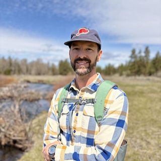 a man in a hat and plaid shirt smiles at the camera with his arms crossed.