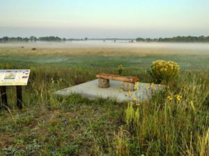 Landscape view overlooking a wide, flat prairie with a layer of fog over it in the distance and a wood-and-stone bench and interpretive sign in the foreground.
