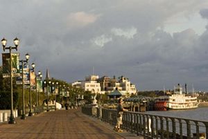 A man looks out over the water along a riverwalk with a riverboat in the background at sunrise.