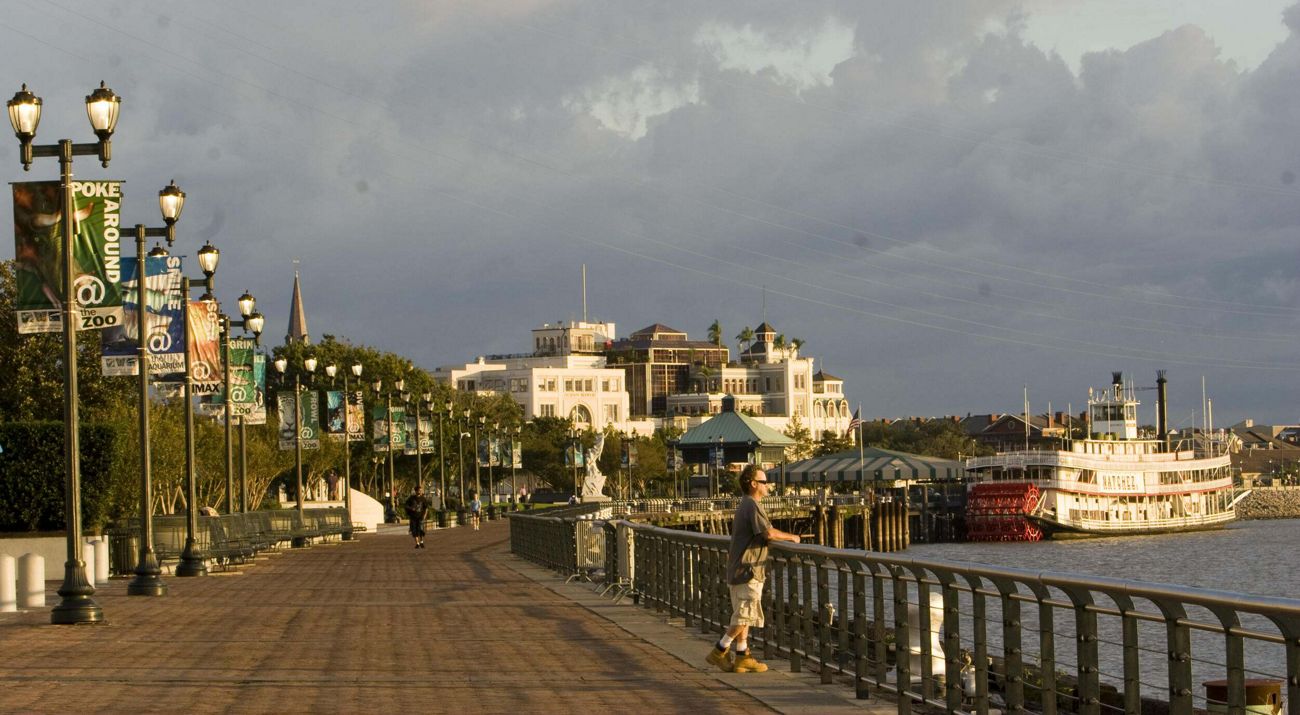 A man looks out over the water along a riverwalk with a riverboat in the background at sunrise.
