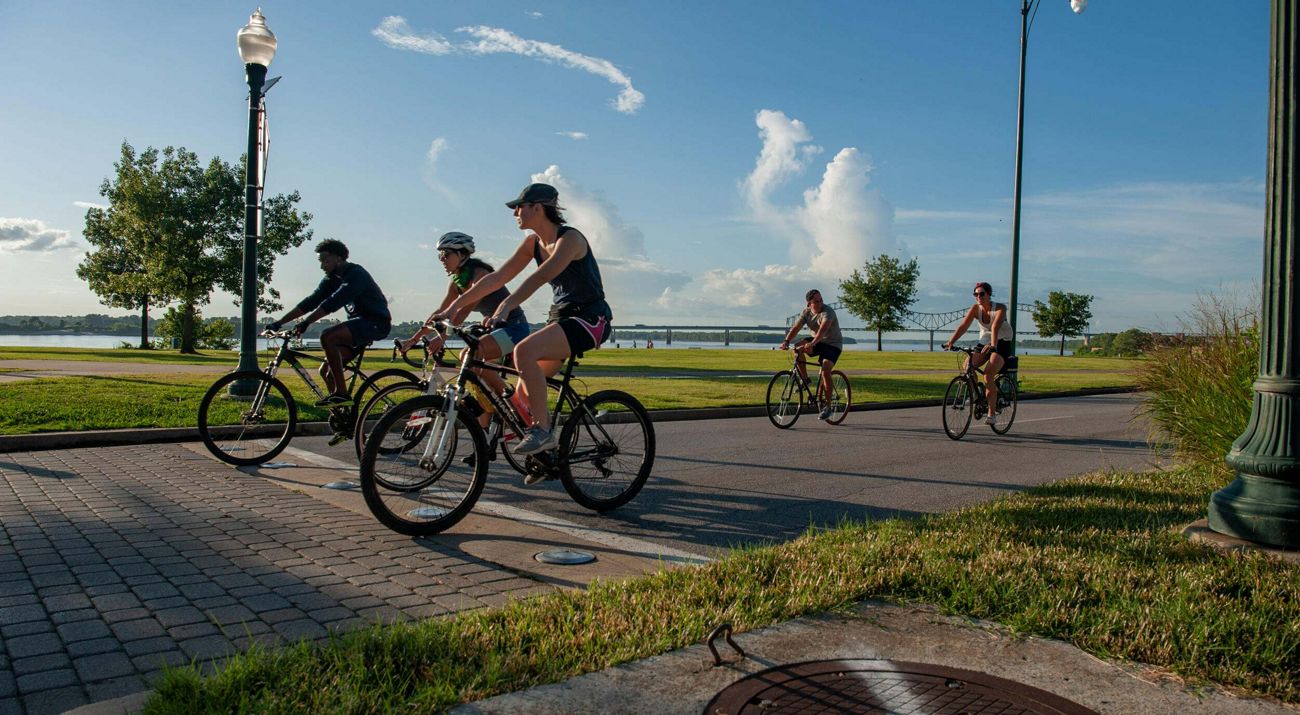 Bicyclists ride on a street in a park setting alongside a river with a bridge over the river in the background.