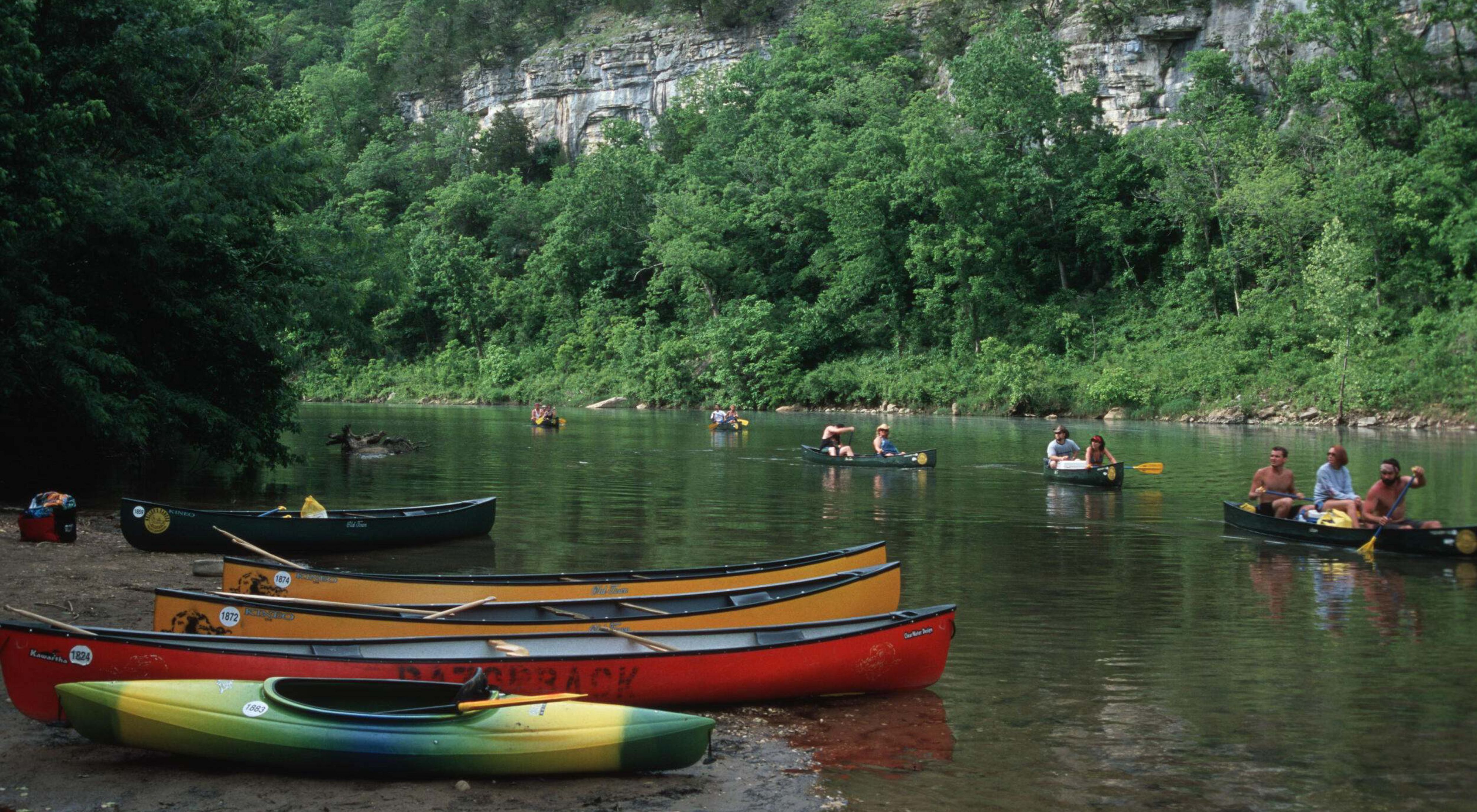 Canoeists on a river lined by trees and a cliff.
