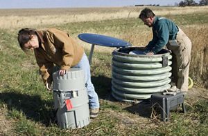 Two works inspect drum-like water monitoring devices in the middle of a farm field.