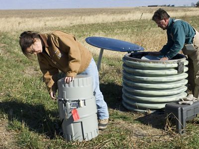Staff members check drum-like water monitoring devices in the middle of a farmfield.