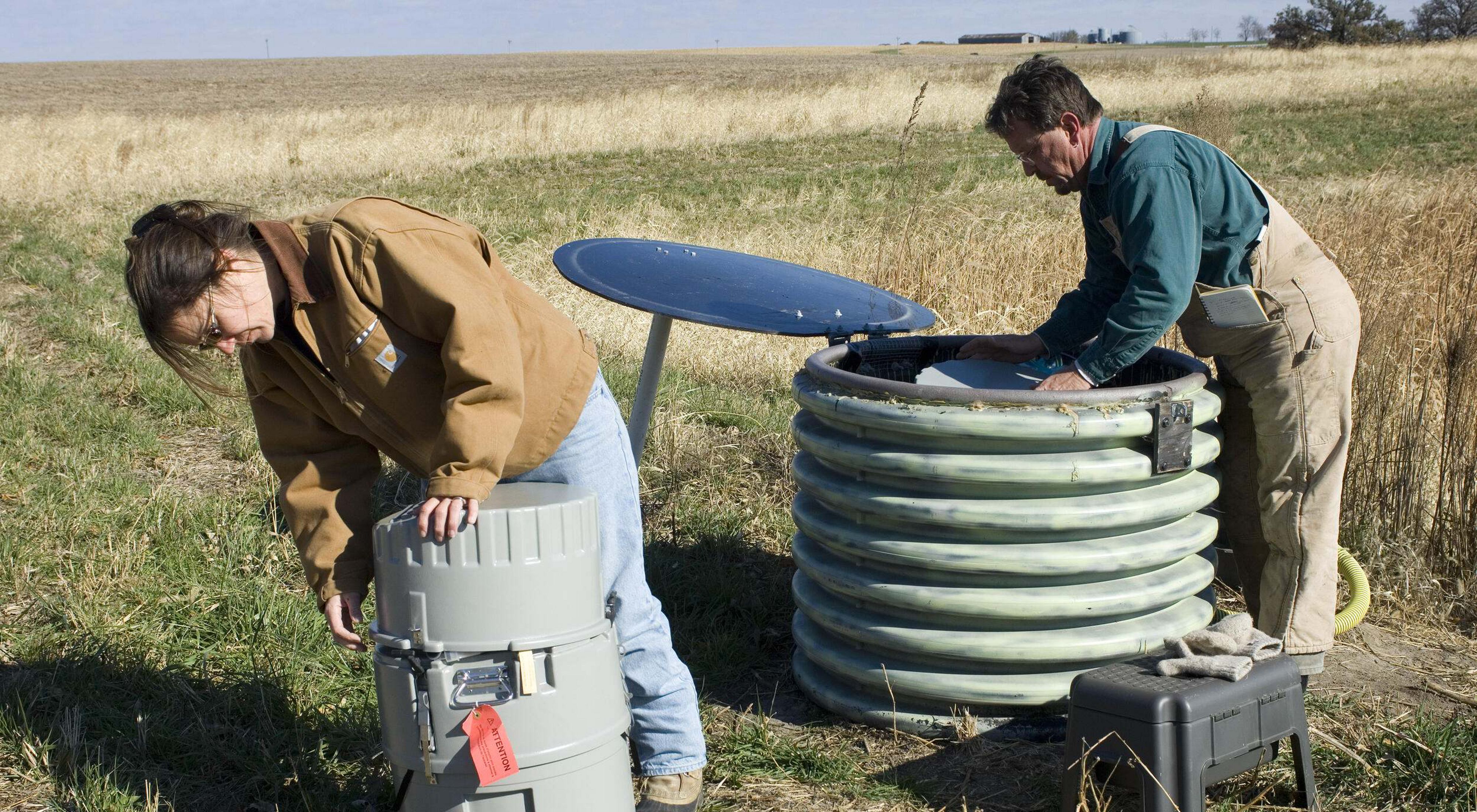 Two works inspect drum-like water monitoring devices in the middle of a farm field.