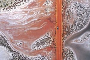 Aerial view looking down at a car driving on a dirt road through desert terrain.