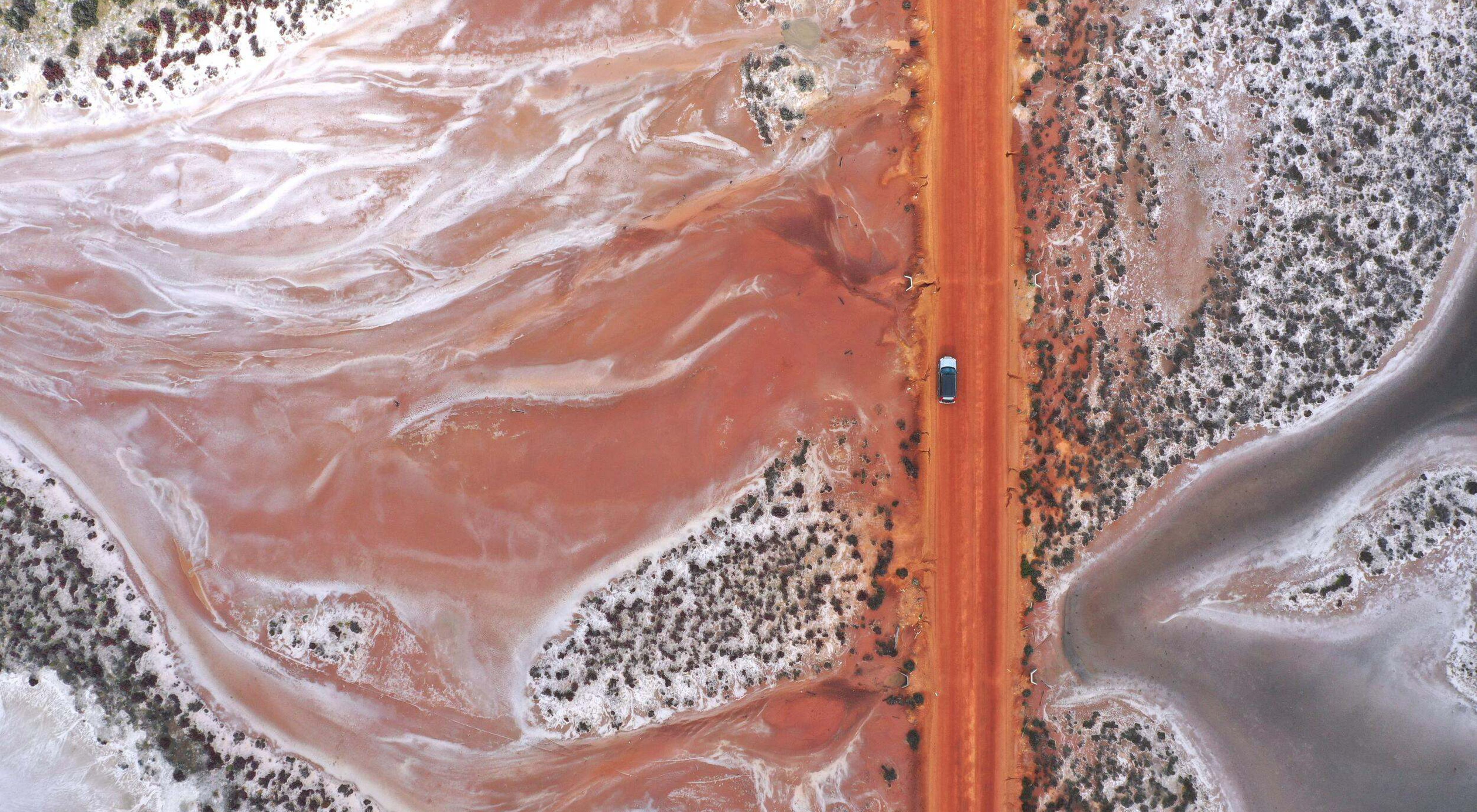 Aerial view looking down at a car driving on a dirt road through desert terrain.