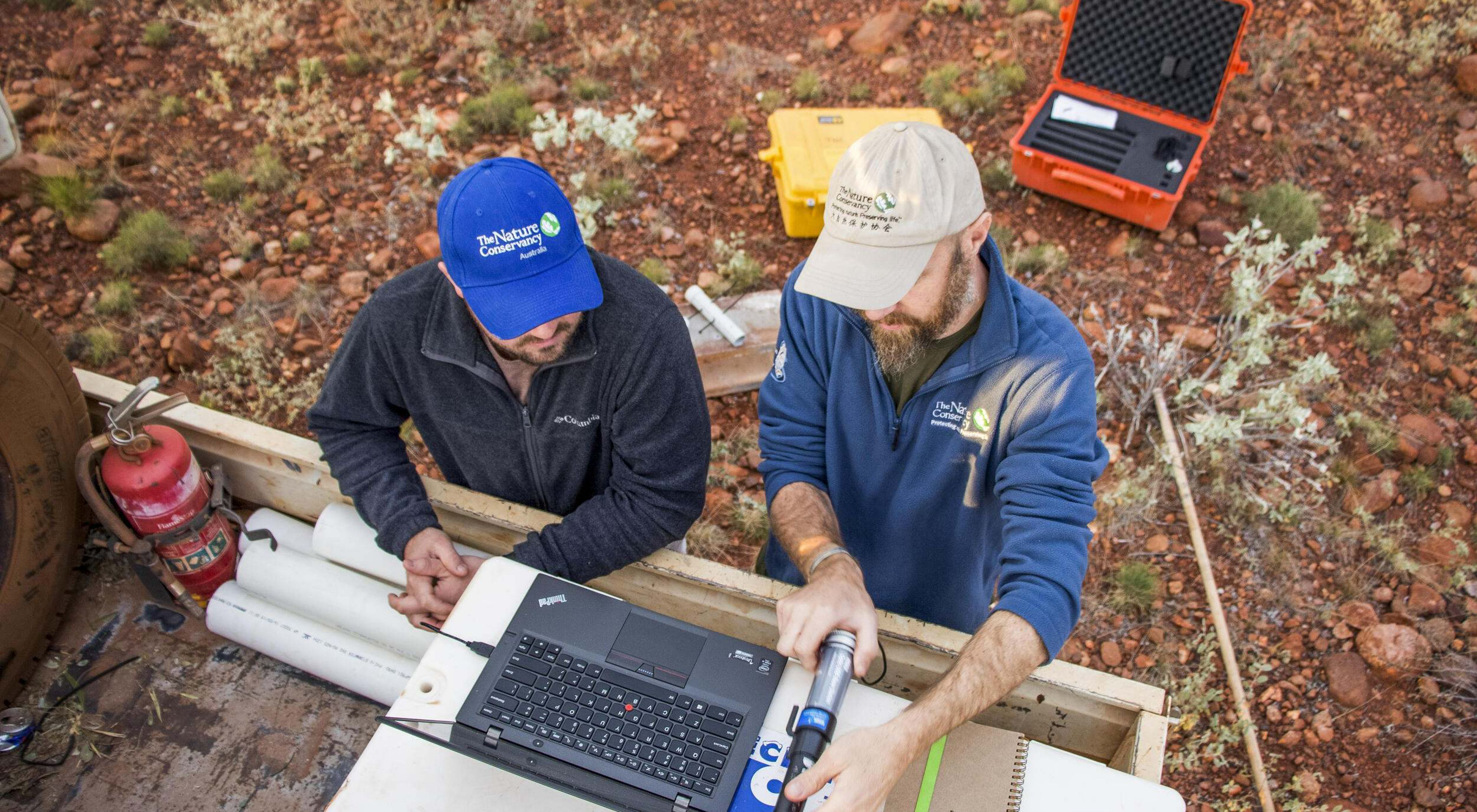 Two staff members wearing TNC hats outdoors while examining data on a laptop computer.
