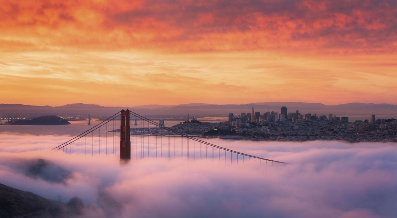 One of the towers of the Golden Gate Bridge pierces through a layer of low fog at sunrise with the San Francisco skyline and a ridge of hills in the background.