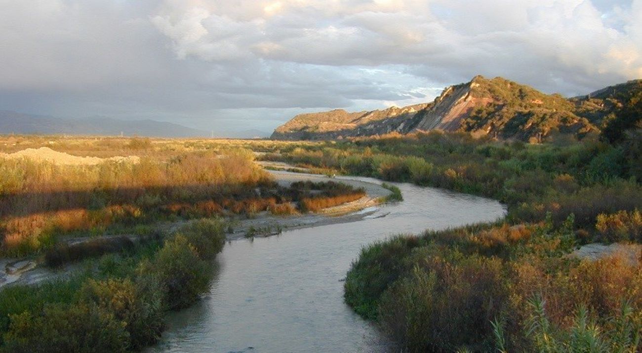 A river winds alongside a plain on one side and a rugged bluff on the other.