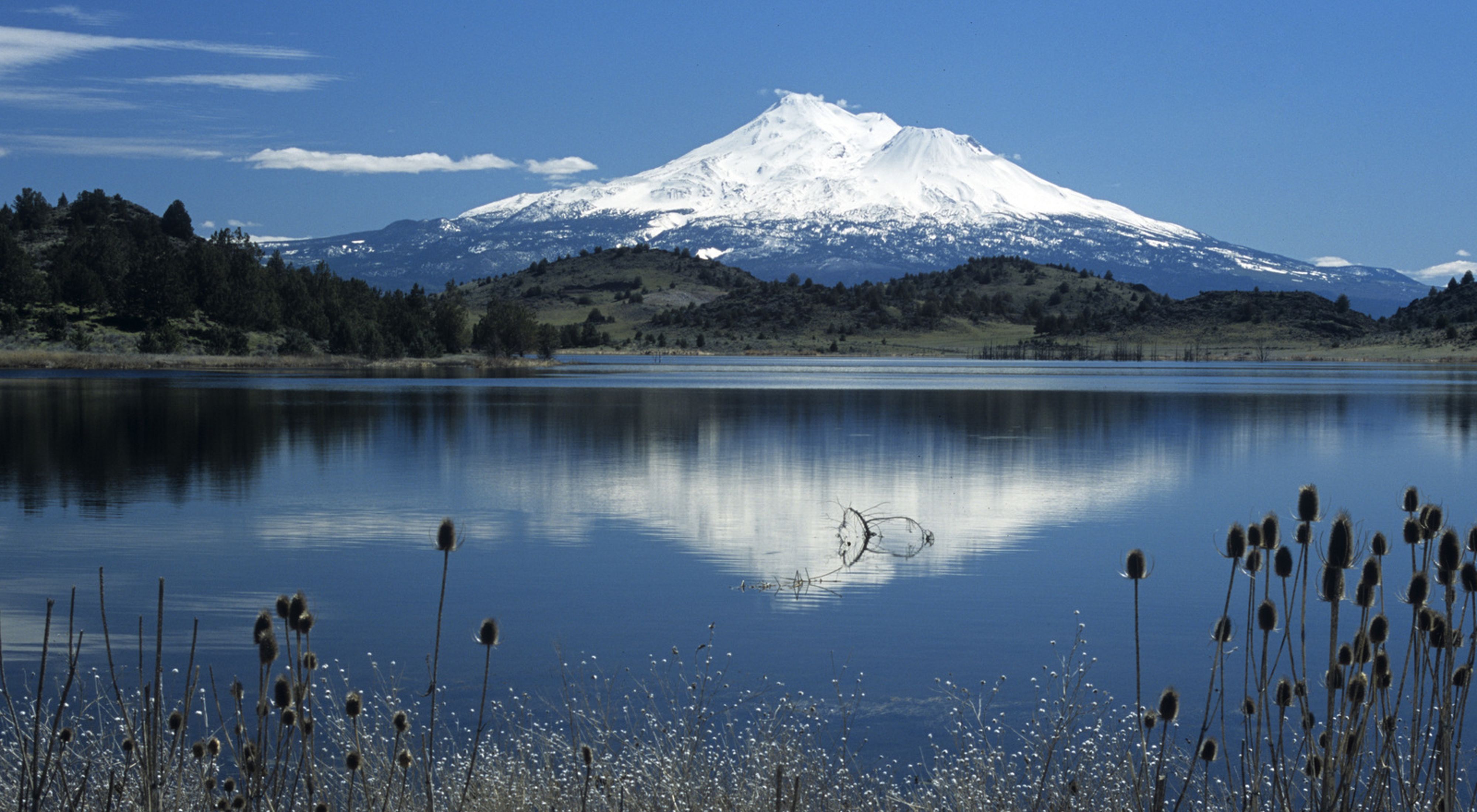Snowcapped volcanic peak in the distance, reflected in the waters of a lake in the foreground.
