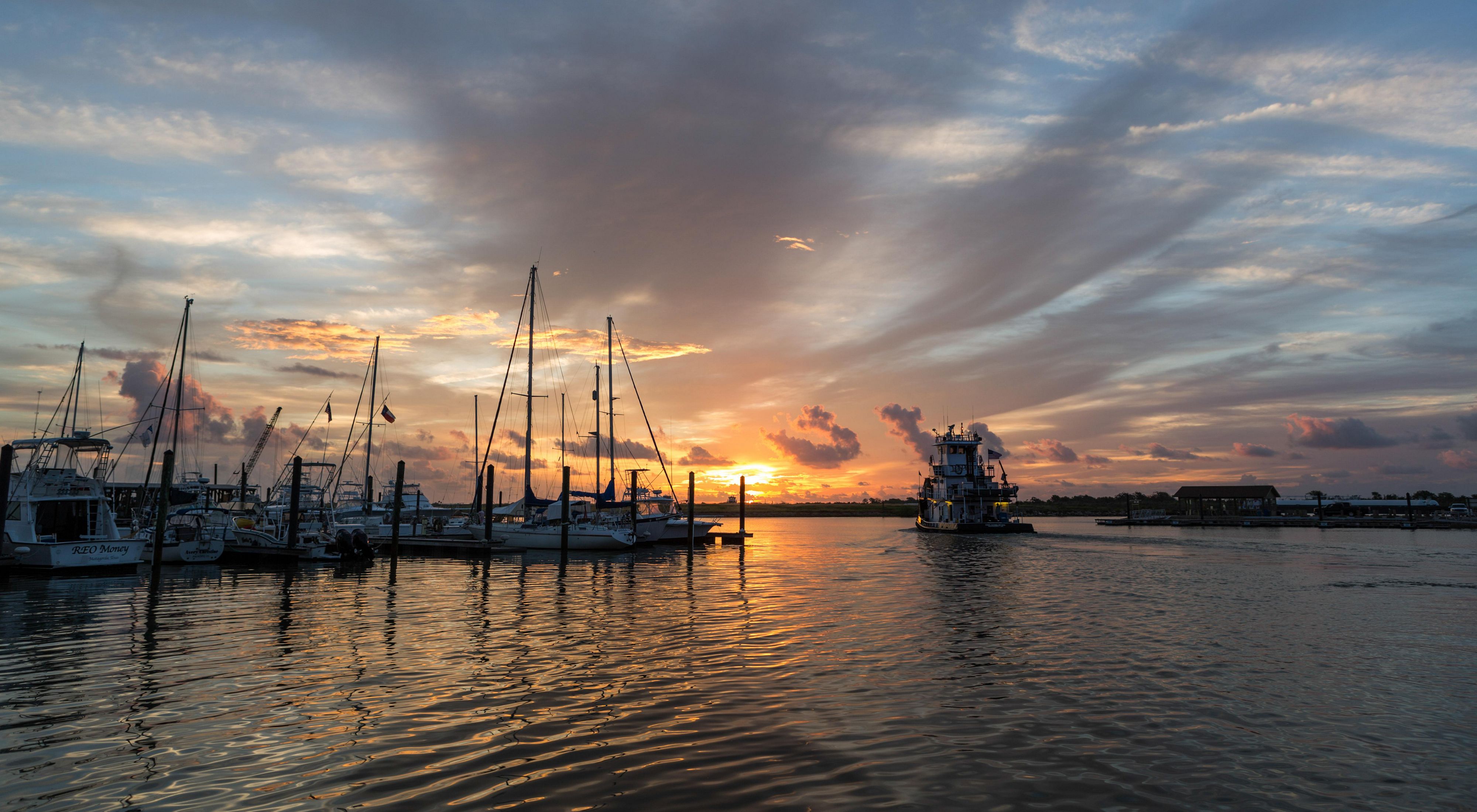 A marina full of sailboats with an orange and pink sunset behind it in a cloud-streaked sky.