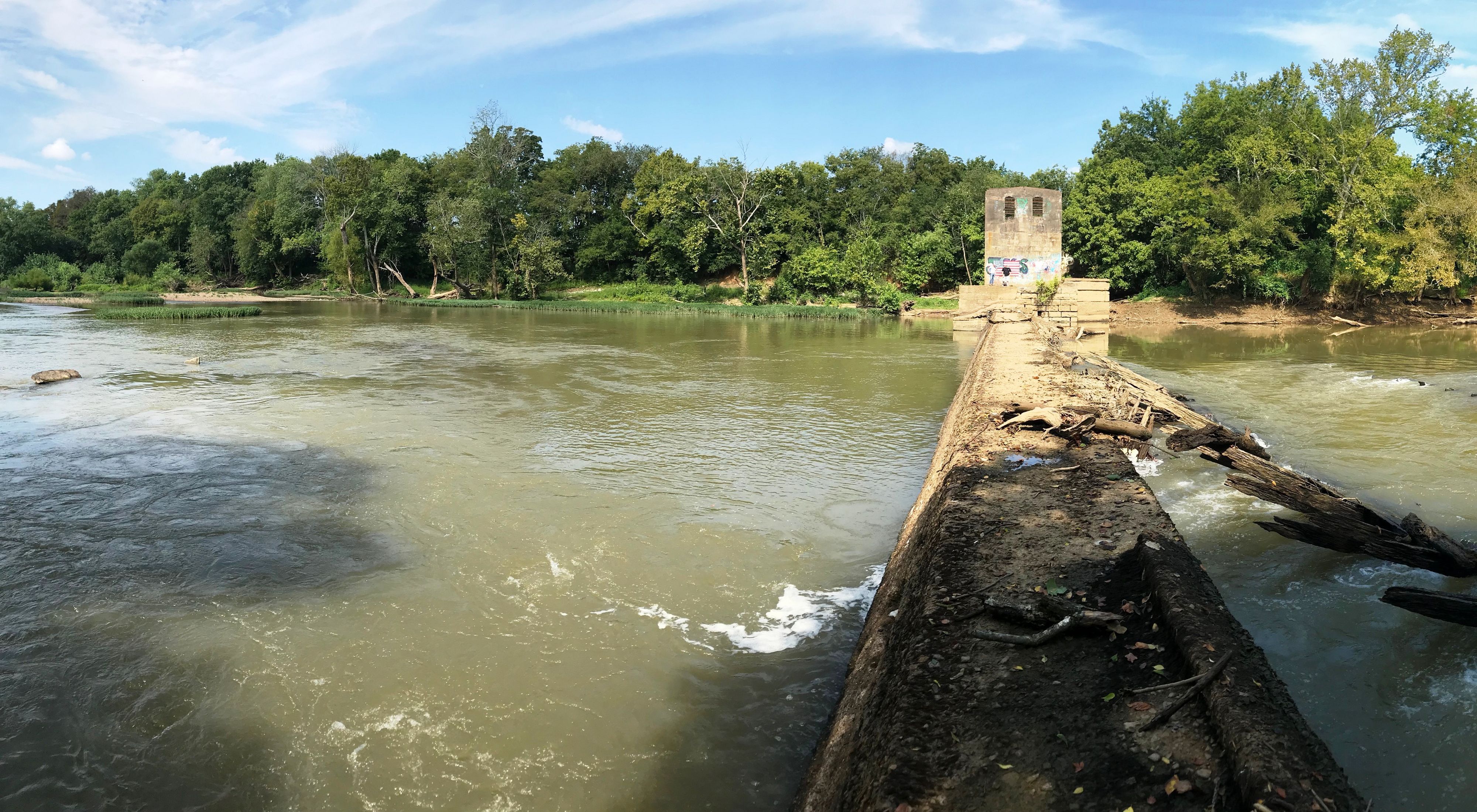 A low dam extends from the camera away across a muddy river, with driftwood piled up on the upstream side of the dam.