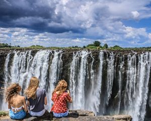 Three young people face Victoria Falls while their hair blows in the wind.