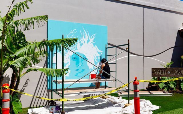 An artist sits on scaffolding painting a mural of a woman with marine organisms for hair and a bright turquoise background.